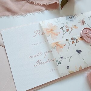 May include: A floral invitation suite featuring a light pink envelope and a card with the text "will you Brides". The invitation has a floral pattern with peach and blue flowers, and a pink wax seal.