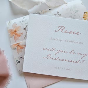 May include: A blush-colored invitation with the name "Rosie" and the text "I can't say 'I do' without you, will you be my Bridesmaid?" is displayed. The invitation is on textured paper, with a floral envelope and ribbon.