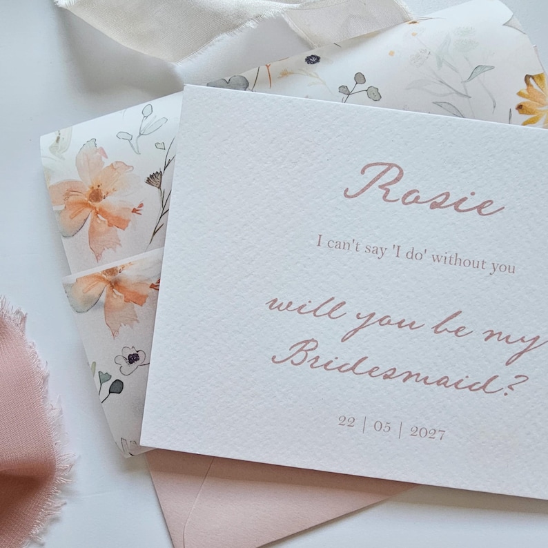 May include: A blush-colored invitation with the name "Rosie" and the text "I can't say 'I do' without you, will you be my Bridesmaid?" is displayed. The invitation is on textured paper, with a floral envelope and ribbon.