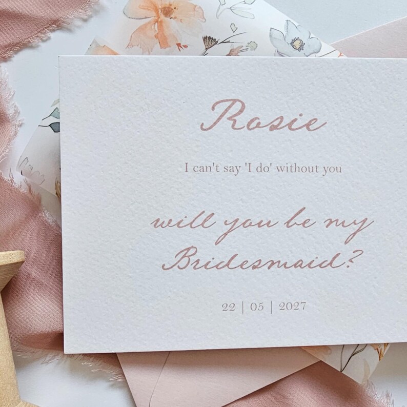 May include: A light pink invitation card with the name "Rosie" in elegant script. The text reads "I can't say 'I do' without you" and "will you be my Bridesmaid?" with the date "22|05|2027" at the bottom. Floral patterned paper is visible in the background.