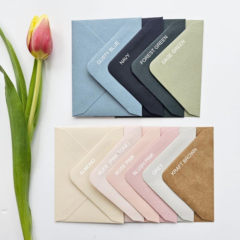 May include: An assortment of colorful envelopes in various shades, including dusty blue, navy, forest green, and sage green. Additional colors are almond, nude, rose pink, blush pink, grey, and kraft brown. A tulip with pink and yellow petals is in the image.