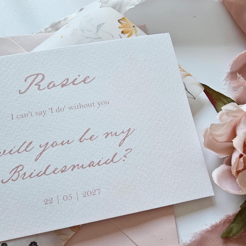 May include: A light pink invitation card with the name "Rosie" in elegant script. The text "I can't say 'I do' without you" and "Will you be my Bridesmaid?" are also printed on the card, along with the date 22/05/2027.