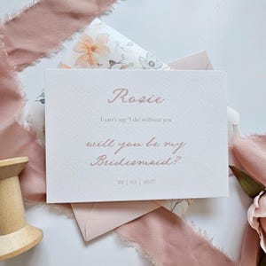 May include: A blush-colored invitation card with the name "Rosie" in elegant script. The text reads "I can't say 'I do' without you, will you be my Bridesmaid?" with the date 22|05|2027. The card is on a floral patterned background.