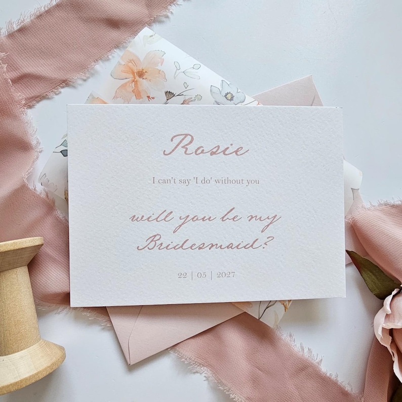 May include: A blush-colored invitation card with the name "Rosie" in elegant script. The text reads "I can't say 'I do' without you, will you be my Bridesmaid?" with the date 22|05|2027. The card is on a floral patterned background.