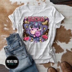 Op de afbeelding: Wit T-shirt met een kleurrijke cartoon stier en de tekst "SUCKA for love". De stier heeft paarse vacht, een roze neus en likt aan een lolly. Het shirt wordt gecombineerd met een blauwe jeans.
