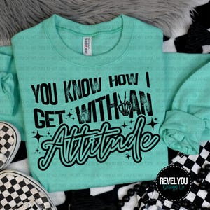 May include: A light teal sweatshirt with the text "YOU KNOW HOW I GET WITH AN Attitude" in black. The design includes a peace sign made of bones and stars. The sweatshirt is displayed with checkered sneakers.