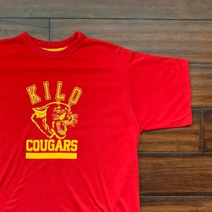 May include: Red t-shirt with yellow trim and a yellow graphic of a cougar with the text "KILO COUGARS".