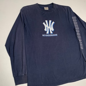Puede incluir: Una camiseta de manga larga azul marino con un gráfico blanco y azul del logotipo de los New York Yankees y la palabra "Neighborhood" debajo. El texto "Neighborhood" se repite verticalmente en la manga derecha.