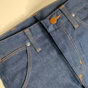 May include: Close-up of the waistband of a pair of blue denim jeans with a brown button and stitching. The jeans have a classic five-pocket style.