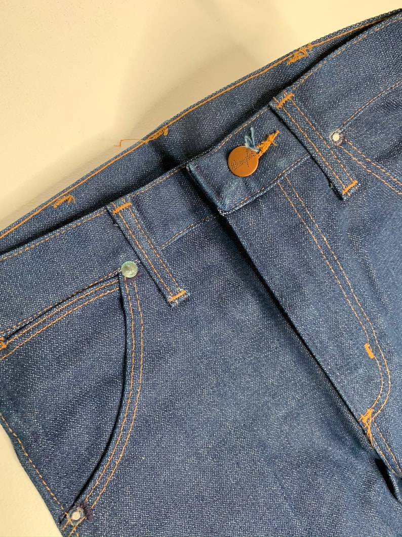 May include: Close-up of the waistband of a pair of blue denim jeans with a brown button and stitching. The jeans have a classic five-pocket style.