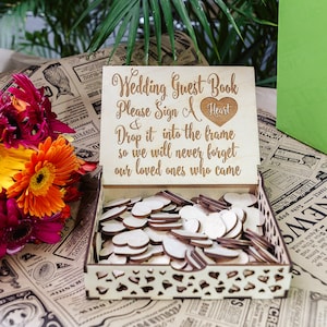 Puede incluir: Una caja de madera con un letrero que dice "Wedding Guest Book Please Sign A Heart & Drop it into the frame so we will never forget our loved ones who came". La caja está llena de pequeños corazones de madera.