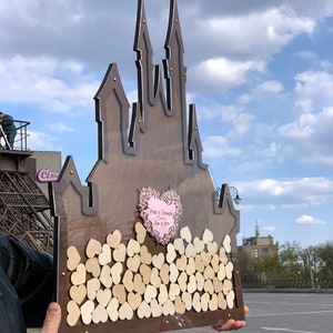 May include: A wooden castle-shaped guest book with a clear acrylic front. The castle is decorated with small wooden hearts. The front of the castle has a pink heart with the text "Craig & Jennifer June 8, 2019".