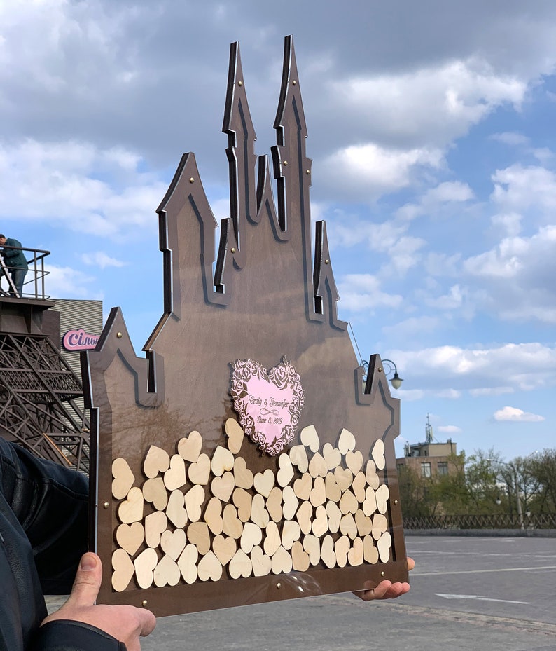 May include: A wooden castle-shaped guest book with a clear acrylic front. The castle is decorated with small wooden hearts. The front of the castle has a pink heart with the text "Craig & Jennifer June 8, 2019".