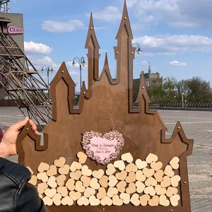 May include: A wooden castle-shaped guest book with a pink heart in the center that says "Craig & Jennifer June 8, 2019". The castle is filled with small wooden hearts.