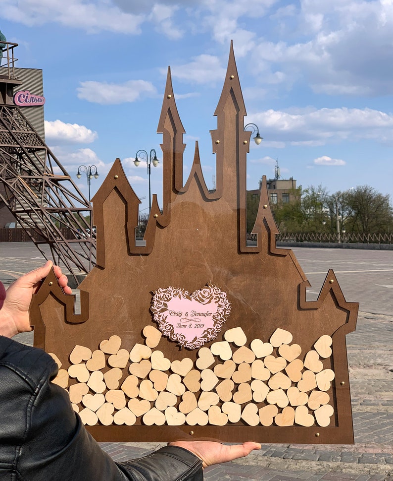 May include: A wooden castle-shaped guest book with a pink heart in the center that says "Craig & Jennifer June 8, 2019". The castle is filled with small wooden hearts.