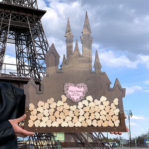 May include: A wooden castle-shaped guest book with a pink heart cutout and the text "Craig & Jennifer June 8, 2019". The castle is filled with small wooden hearts.