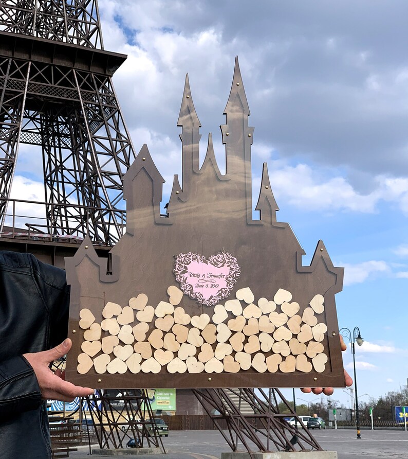 May include: A wooden castle-shaped guest book with a pink heart cutout and the text "Craig & Jennifer June 8, 2019". The castle is filled with small wooden hearts.