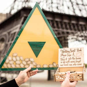 Könnte beinhalten: Eine hölzerne Gästebuchbox mit einem Schild, auf dem steht "Wedding Guest Book Please Sign A Drop it into the frame so we will never forget our loved ones who came". Die Box ist mit kleinen, diamantförmigen Holzstücken gefüllt. Die Box wird von einer Hand gehalten. Im Hintergrund ist eine große Metallstruktur zu sehen.