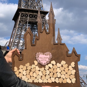 May include: A wooden castle-shaped guest book with a pink heart that says "Craig & Jennifer June 8, 2019". The castle is filled with small wooden hearts.