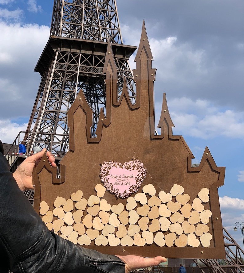 May include: A wooden castle-shaped guest book with a pink heart that says "Craig & Jennifer June 8, 2019". The castle is filled with small wooden hearts.