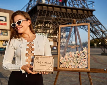 May include: A woman in a white top and sunglasses holds a wooden box with the inscription "Emily & Michael 10.12.23."  The box is in front of a wooden easel with a glass frame filled with small wooden hearts. The easel is in front of a replica of the Eiffel Tower.