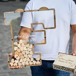 May include: Wooden guest book alternative in the shape of the letter "I" with a clear front, filled with small wooden hearts. The frame has the names "Art & Jamie" and the date "October 10, 2025" etched on it. A matching wooden box is also included.