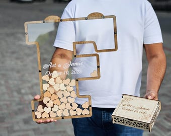 May include: Wooden guest book alternative in the shape of the letter "I" with a clear front, filled with small wooden hearts. The frame has the names "Art & Jamie" and the date "October 10, 2025" etched on it. A matching wooden box is also included.