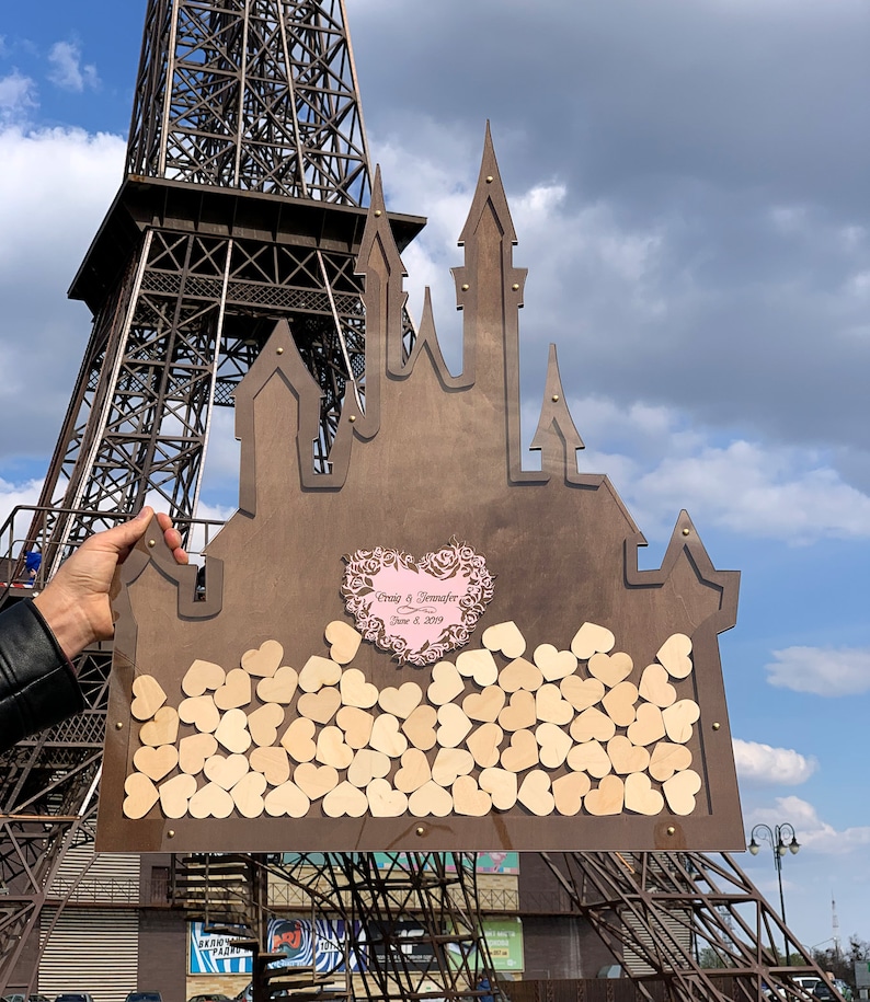 May include: A wooden castle-shaped guest book with a pink heart and the text "Craig & Jennifer June 8, 2019". The castle is filled with small wooden hearts.