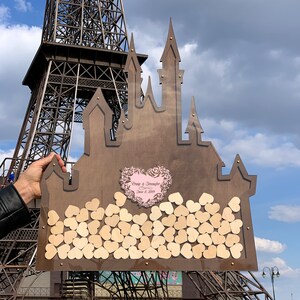 May include: A wooden castle-shaped wedding guest book with a heart-shaped cutout for guests to sign their well wishes. The heart cutout is decorated with a pink floral design and the text "Craig & Jennifer June 8, 2019".