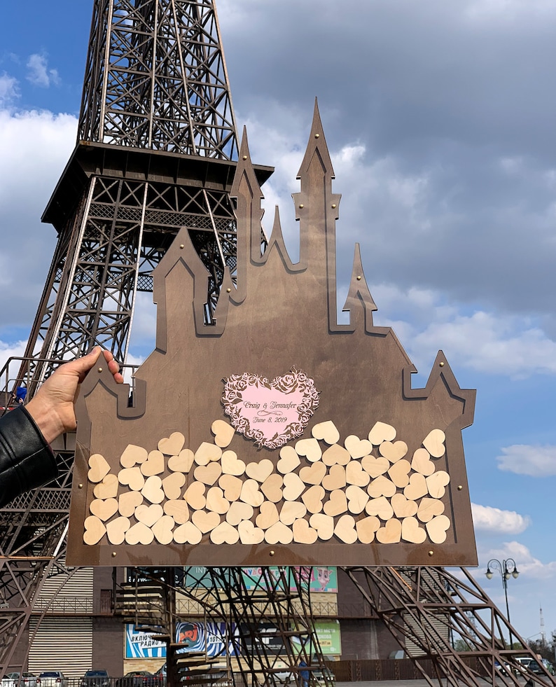 May include: A wooden castle-shaped wedding guest book with a heart-shaped cutout for guests to sign their well wishes. The heart cutout is decorated with a pink floral design and the text "Craig & Jennifer June 8, 2019".