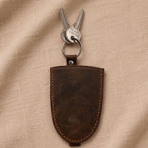 May include: A dark brown leather key holder with a shield-like shape and visible stitching along the edges. A silver-toned metal ring is attached to the top, holding two silver-colored metal keys. The leather has a matte, textured finish.