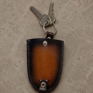 May include: A shield-shaped leather key holder with a gradient finish transitioning from dark brown edges to a tan center. It features a silver metal snap button closure at the top and bottom, a metal key ring, and two silver metal keys attached.