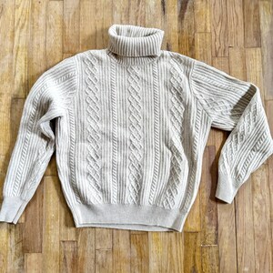 May include: A beige turtleneck sweater with a cable knit design. The sweater has a ribbed collar and cuffs, and the body of the sweater features a textured pattern. The sleeves are long and the sweater is made of a soft material.
