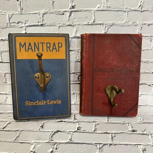 May include: Two vintage book-shaped wall hooks. One is a blue book with "MANTRAP" and "Sinclair Lewis" text, the other is a red book with "ENGLISH GRAMMAR" text. Both have bronze-colored hooks.