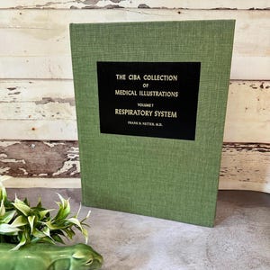 Könnte beinhalten: Ein grünes Hardcover-Buch mit dem Titel The Ciba Collection of Medical Illustrations Volume 7 Respiratory System von Frank H. Netter, M.D. Der Einband zeigt ein schwarzes rechteckiges Etikett mit goldenem Text auf einem strukturierten grünen Stoff.