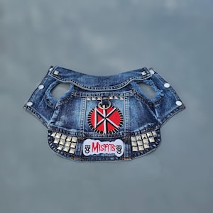 Op de afbeelding: Een blauw denim hondenvest met een distressed look, met een rood en zwart embleem met een symbool en het woord "Misfits". Het vest is versierd met zilveren studs langs de onderkant en rond de armsgaten.