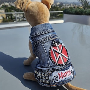 May include: A plush dog toy wearing a denim vest with silver studs and patches. The patches include a red and white design and the word "MISFITS" in red lettering. The dog is tan and sitting on a gray surface.