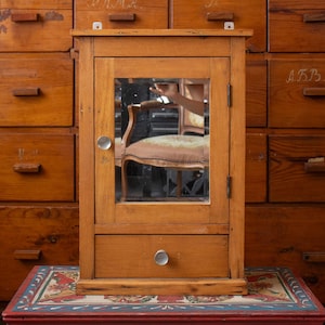 Puede incluir: Un armario de pared rectangular de madera con una puerta única que presenta un panel de espejo central y un pequeño pomo de tono plateado. Debajo de la puerta hay un cajón horizontal único con un pomo de tono plateado a juego.