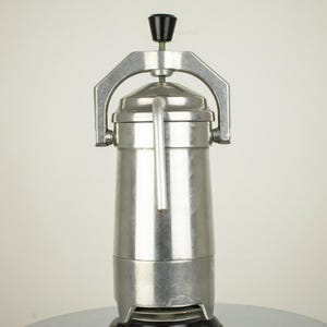 May include: A vintage, silver-toned espresso maker with a black handle and base. The coffee maker has a cylindrical body, a curved handle, and a pressure release valve. It is set on a shiny, chrome-like surface.