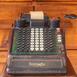 Pode incluir: Uma máquina de somar Burroughs vintage com caixa de metal escuro, placa frontal verde e uma manivela manual no lado direito. O dispositivo possui várias colunas de teclas circulares com números brancos e um suporte para rolo de papel.