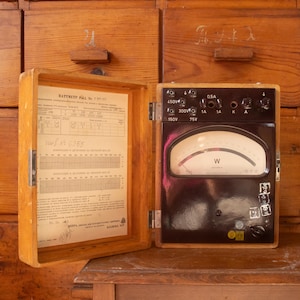 Op de afbeelding: Een vintage wattmeter in een houten behuizing met een scharnierend deksel. De binnenkant van het deksel bevat een technische tabel met Cyrillische tekst. Het apparaat heeft een zwart front met een gebogen analoge meter, meerdere aansluitklemmen en labels waaronder 450V, 300V, 150V, 0,5A, 1A, K en A.