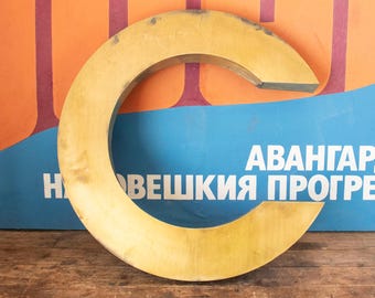 Può includere: Una grande lettera C in legno dorato. La lettera è appoggiata su una superficie di legno. La lettera è davanti a un poster blu e arancione con il testo "АВАНГАРД НОВЕШКИЙ ПРОГРЕСС".
