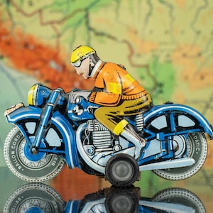 Może przedstawiać: Zabytkowy motocykl zabawkowy w kolorze niebieskim i żółtym z jeźdźcem w kolorze pomarańczowym i żółtym. Motocykl ma szczegółowy design ze słowami "BALLON CORDA" na kołach. Tło to mapa.
