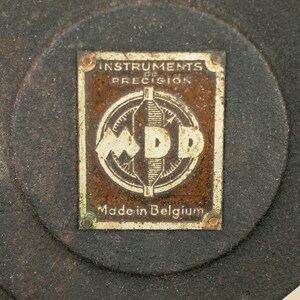May include: A close-up of a circular, metal measuring tool with a rectangular, brass plaque. The plaque reads "Instruments de Precision" and "Made in Belgium" with a logo of "MDP" in the center.