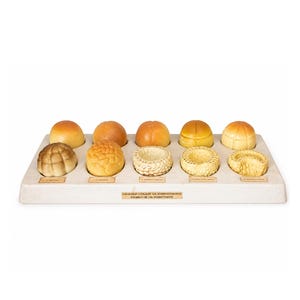 Könnte beinhalten: Eine Präsentation von Brötchen in verschiedenen Entwicklungsstadien. Die Brötchen reichen von hellbraun bis goldbraun und weisen unterschiedliche Texturen auf. Die Präsentation befindet sich auf einem rechteckigen, cremefarbenen Tablett.