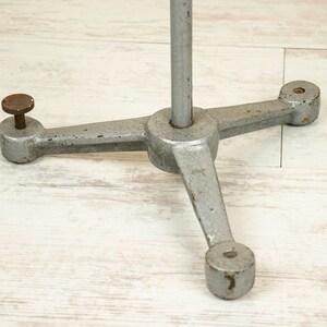 May include: A silver metal tripod stand with three legs and a round base. The stand is designed to hold a light or other object.