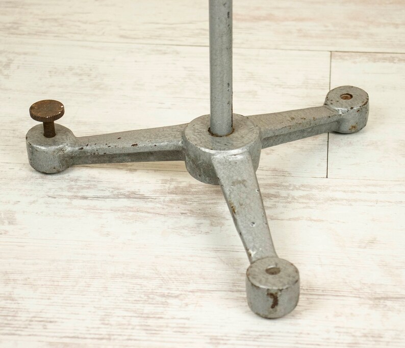 May include: A silver metal tripod stand with three legs and a round base. The stand is designed to hold a light or other object.