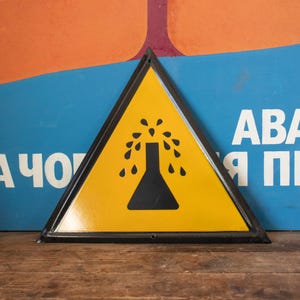 May include: Yellow triangular warning sign with a black border. The sign has a black silhouette of a beaker with liquid pouring out of it. The sign reads "ABA" and "Я П"