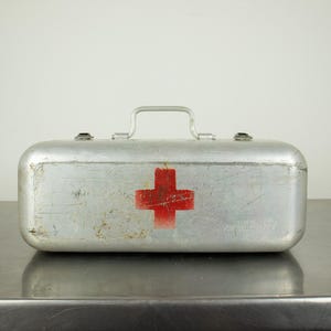 May include: A rectangular silver-coloured metal first aid kit featuring a prominent red cross symbol on the front. The container has a top-mounted metal handle and two metal latches. The surface shows signs of wear, including scratches and scuffs.