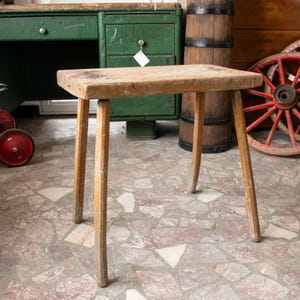 May include: A small, rustic wooden stool with a rectangular seat and four angled legs. The wood has a natural, light brown colour with visible grain and signs of wear. The stool is approximately 46 cm tall.